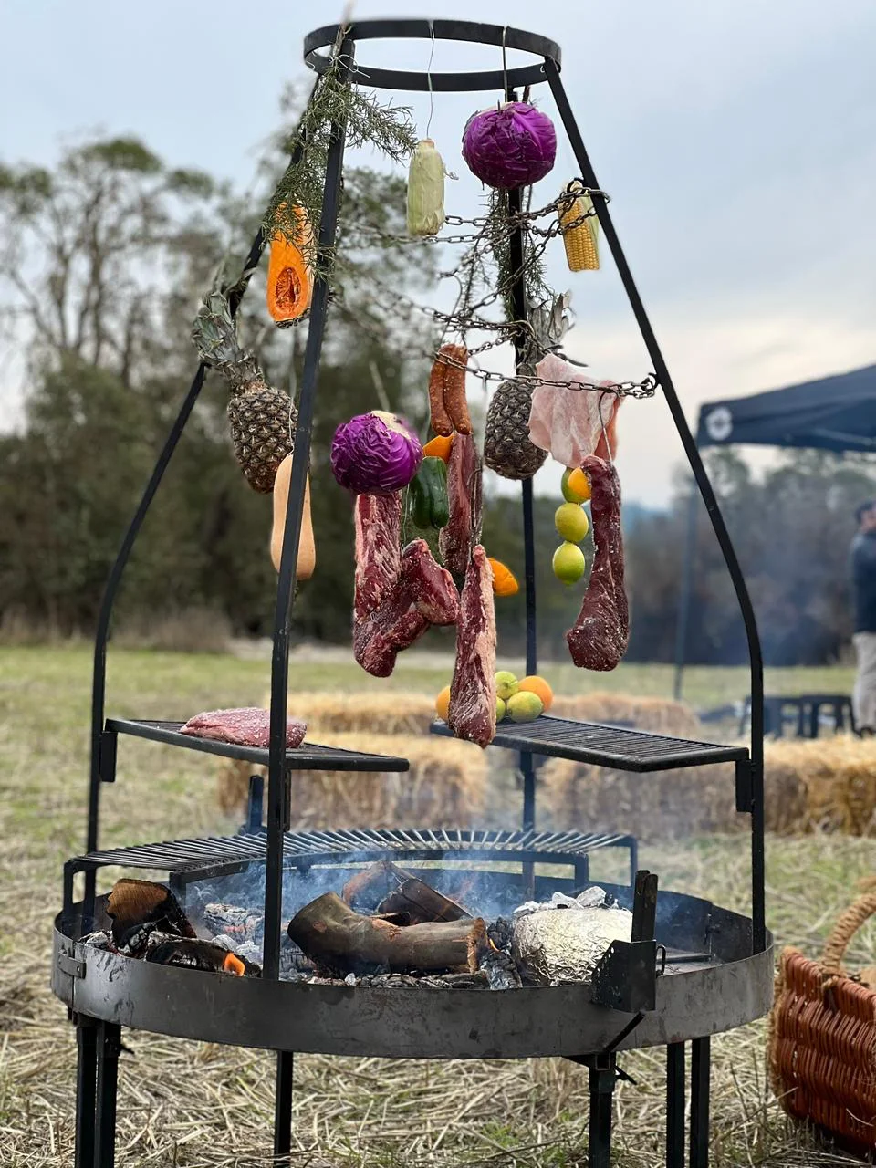 Cruz de fierro con cortes de carne, embutidos artesanales, frutas y flores junto a un fogón en un asado al aire libre en el campo
