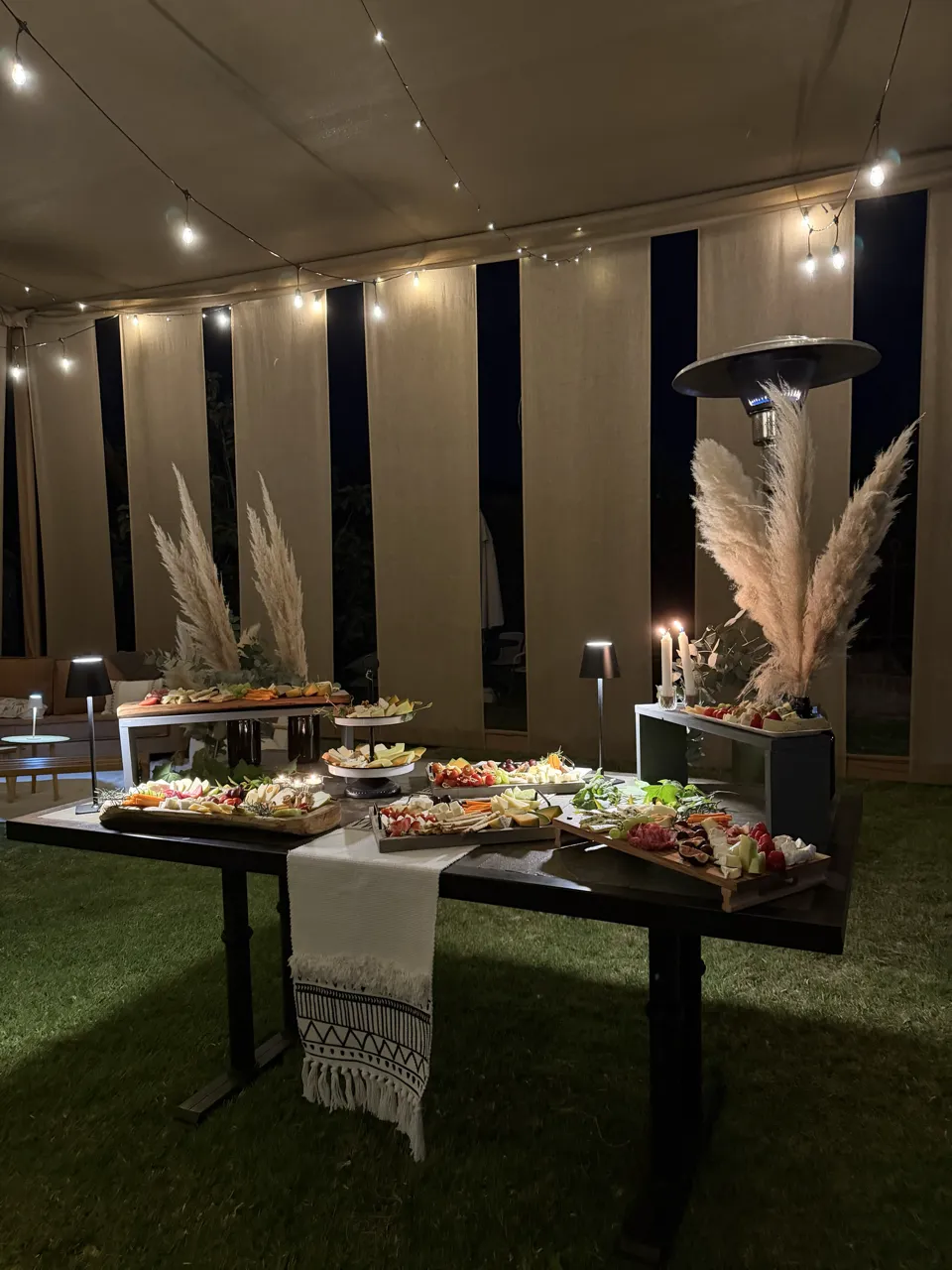 Mesa de tablas de quesos y fiambres iluminada con guirnaldas de luces durante un evento nocturno bajo carpa