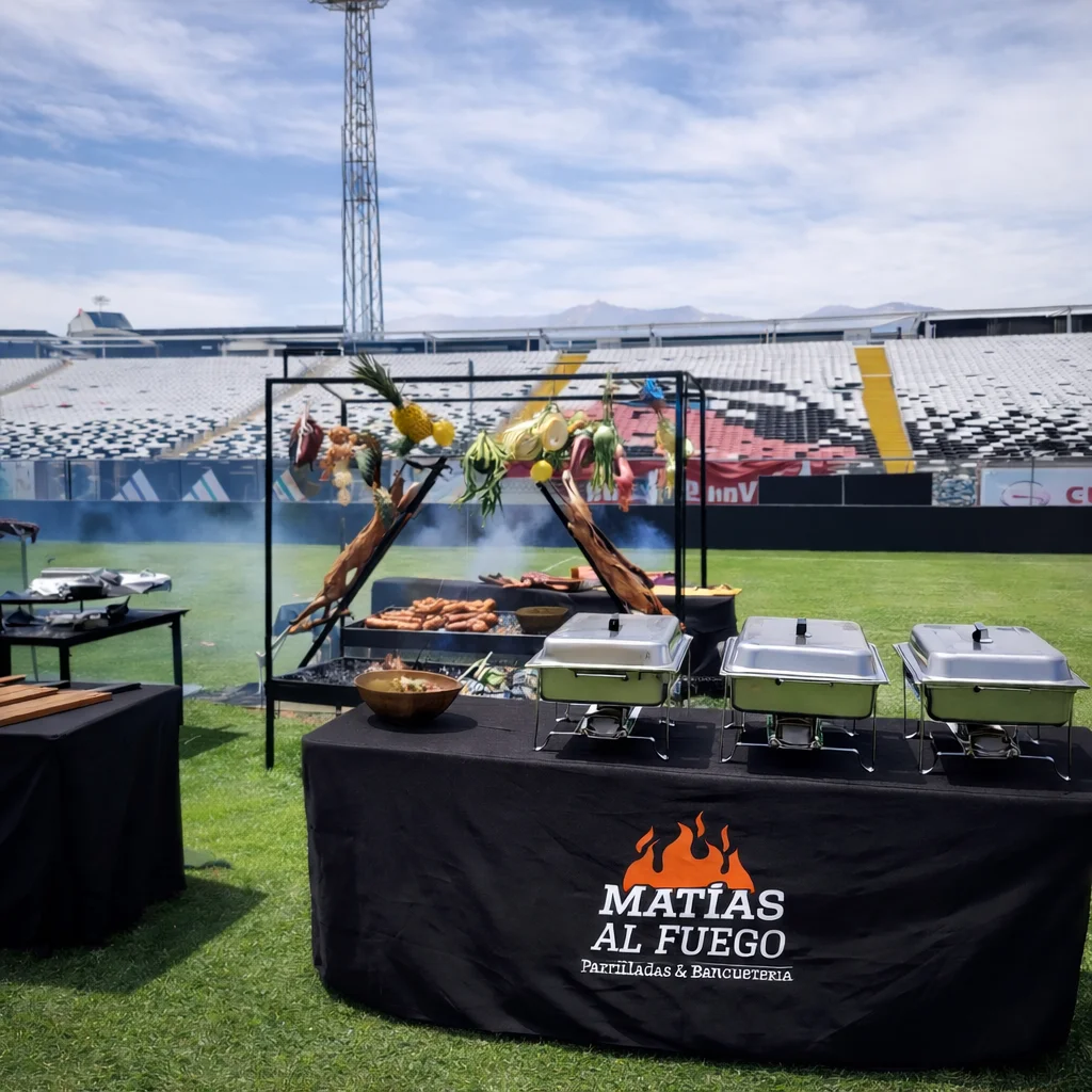 Montaje de banquetería con asado en vivo y mesas con mantel Matías al Fuego en el Estadio Monumental — catering para evento de empresa en Santiago